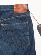 Close-up of Giacomo 77 Dark Used Jeans in mid blue denim with a brown leather patch embossed with Blue de Gênes near the waistband. A string-attached tag hangs from the belt loop, highlighting back pocket details and relaxed loose fit for ultimate comfort.