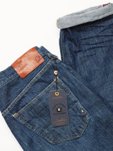 Close-up of Blue de Gênes Giacomo 77 Dark Used Jeans in mid blue rigid denim with a brown leather brand patch on the waistband and a tag adorned with a small red, white, and black shield logo. The relaxed loose fit jeans are folded at the hem, highlighting the fabrics cuff details.