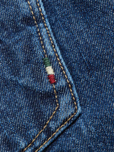 Close-up of Giacomo 77 Dark Used Jeans by Blue de Gênes, showing mid blue denim fabric with a diagonal seam. Near the seam, green, white, and red stitching creates a mini flag stripe. The rigid texture and classic weave highlight its rich color.
