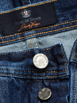Close-up of Giacomo 77 Dark Used Jeans in Mid Blue Denim reveals visible fabric texture. Features black label and metallic button engraved with Blue de Gênes brand name and logo. Rigid denim provides a relaxed loose fit, highlighting detailed stitching.