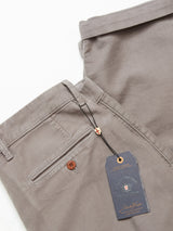The Paco Papago Chino by Blue de Gênes in stormy grey is showcased, featuring a refined style with a buttoned back pocket. A black branded tag is attached to the belt loop. The garment-dyed pants are neatly folded against a white background.