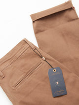 The Menzo Dobby Chino - Brownie by Blue de Gênes features Tuscan cotton for a vintage look, back pocket details, and a stitched emblem. It has a navy blue tag with gold lettering and a coat of arms, while comfort stretch fabric ensures an impeccable fit.