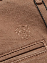 Close-up of Menzo Dobby Chino - Brownie by Blue de Gênes, showcasing comfort stretch brown fabric with a stitched shield-like emblem and visible stitching details on a small pocket, accentuating its vintage appeal.