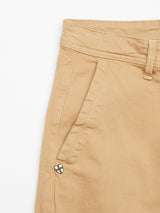 The Menzo Dobby Chino - Khaki by Blue de Gênes features a partial view of the waistband and pocket, showcasing visible stitching and a belt loop. A small black cross emblem on white stands out near the pocket, while comfort stretch fabric offers a perfect vintage look.