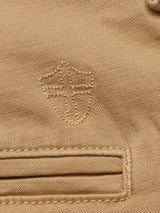 A close-up of the Blue de Gênes Menzo Dobby Chino in Khaki reveals an embroidered emblem of a shield and cross, complemented by a thick-stitched pocket. The fabric offers a textured vintage look with comfort stretch for enhanced wearability.
