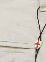 A close-up features a stone Menzo Dobby Chino by Blue de Gênes with a subtle embroidered cross crest. A small pendant, red cross on white, hangs from a black cord, resting on vintage-look fabric enhanced by comfort stretch.