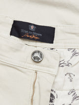 Close-up of Menzo Dobby Chino - Stone by Blue de Gênes, showcasing a vintage style with a silver emblem button. The lining has a monochrome design with cursive writing and small illustrations, providing comfort stretch for easy wear.