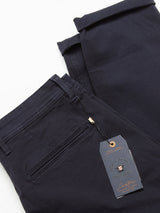 Close-up of a neatly folded pair of Menzo Dobby Chino - Dark Navy by Blue de Gênes, visible tag with text and gold accent. These comfort stretch chinos with a back pocket, on white surface, offer a vintage-inspired look that blends style and functionality.