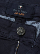 Close-up of Menzo Dobby Chino pants in Dark Navy from Blue de Gênes, featuring comfort stretch fabric and an engraved button. The label showcases similar branding with decorative stitching on partially visible gray denim for a vintage-inspired look.