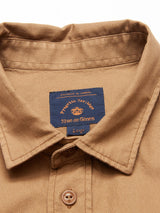 Close-up of a tan cotton piqué button-up shirt collar, featuring a dark blue label with gold text: Porselli Heritage, Blue de Gênes. This garment-dyed Enrico Nidos Shirt - Fudge displays one button beneath the collar.