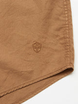 Close-up of the light brown cotton piqué fabric of the Enrico Nidos Shirt - Fudge by Blue de Gênes, featuring a small embroidered shield emblem. The fabric edge is neatly stitched, highlighting the textured pattern typical of this shirt.