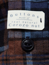 A close-up of the Blue de Gênes Zamboni Quadro Shirt - Navy Check reveals a winter cotton-linen blend with a yarn-dye check pattern in dark brown and navy blue, featuring Corozo nut black buttons, and a beige tag verifying: Buttons made of real natural Corozo nut.