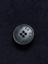 A close-up shows a round, dark buffalo horn button embossed with Blue de Gênes and a small shield emblem on a textured fabric resembling Melton wool, typical of the classic Peacoat - Dark Navy design.