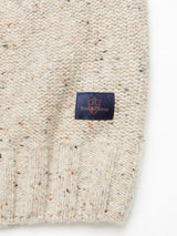 Close-up of the Adamo Knit - Pebble Beige from Blue de Gênes, featuring mélange yarn in beige with a black and orange speckled pattern, plus a small blue patch with an orange shield emblem and Kired Stones near the hem.