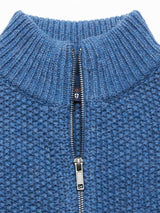 The Aimone Knit Cardigan in Sugar Blue by Blue de GĂȘnes displays a coarse pearl knit pattern with a half-zip design. Partially opened, it reveals the inner lining and logo, with its ribbed collar adding to the intricate weave and classic lambswool charm.