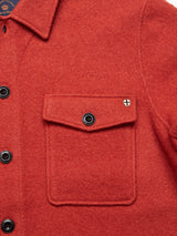 The Pampa Overshirt in Ochre Red by Blue de GĂȘnes features a close-up of its textured boiled wool, black corozo buttons, and a front pocket with a buttoned flap adorned with a small cross-shaped emblem.