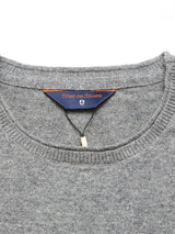 Close-up of a gray crewneck jumper neckline featuring the label Blue de Gênes. The textured Merino wool of the Monte AW Knit - Stormy Grey highlights its quality, with a small string loop and tag adding elegance.