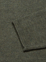 A close-up of Blue de Gênes Monte AW Knit - Forest shows its knit olive green fabric with a visible sleeve. The sweater features slubby yarn, ribbed cuffs, and appears to be made from premium Merino wool.