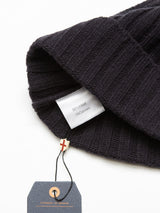 A close-up of the Oscar Croft Beanie in Dark Navy by Blue de Gênes showcases its ribbed texture, fabric tag indicating a 90% wool and 10% cashmere composition, and a dangling tag with a cross symbol on this 5-gauge knit accessory.