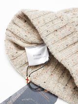 A close-up of the Oscar Melange Beanie in Pebble Beige by Blue de Gênes reveals its ribbed texture and a tag reading 80% Wool, 20% Recycled Nylon. A charm with a red cross hangs from a black string, highlighting its soft wool blend design.