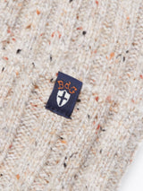 Close-up of the Oscar Melange Beanie in Pebble Beige by Blue de Gênes, featuring a beige knit fabric with colorful speckles and a small black tag adorned with orange stylized letters and a white shield-like emblem.