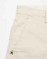Close-up of the natural Abramo Desk Shorts by Blue de Gênes, featuring a waistband and front pocket. A small metal emblem with a cross design is pinned near the pocket. Made from organic cotton, the subtly textured fabric adds to their charm.