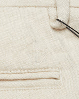 A close-up of beige organic cotton fabric with a visible seam and stitching line shows a black string looped through a top belt loop. The texture, reminiscent of Blue de Gênes Abramo Desk Shorts - Natural, features a small embroidered design near the center.