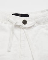 Close-up of Blue de Gênes Crivello Cord Shorts in Snow White showing a drawstring waist, dark button above the zipper, and ribbed waistband with a partially visible logo tag. This garment-dyed piece exudes subtle charm.