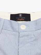 A close-up of the Elak Riga Shorts in blue stripe showcases a fine line cotton waistband with a button and zipper. A black label with Blue de Gênes in white and orange lettering is sewn inside, adding elegance to the relaxed regular fit.