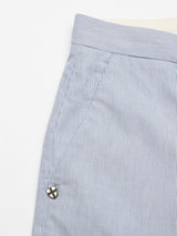 A close-up of Elak Riga Shorts - Blue Stripe from Blue de Gênes showcases relaxed regular fit light blue and white striped pants with an elastic waistband. A small black and white geometric embroidered logo accents the fine line cotton fabric near the bottom of the leg, set against a plain white background.