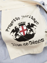 Close-up of a jackets inner lining shows a black emblem with a red cross and two lions. Text reads Blue de Gênes. The relaxed-fit jacket is partially open, revealing striped blue fabric and elegant Corozo nut buttons.