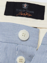 The Elak Riga Shorts by Blue de Gênes feature a detailed black label beside a red cross emblem, crafted from fine line cotton fabric in blue and white stripes. The eco-friendly design includes two Corozo nut buttons, partially covered by beige fabric.