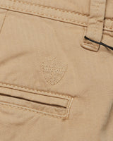 Close-up of Blue de Gênes Menzino Shorts in khaki, featuring a textured shield emblem above the pocket. The garment-dyed fabric displays a herringbone pattern and includes a visible belt loop at the top right.