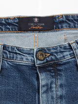 A close-up of the Repi Roll Jeans - Mid Blue Denim by Blue de Gênes shows the blue denim waistband with a black label, a metallic button with an engraved logo, and distinct orange stitching on belt loops and seams. Made from comfort stretch denim for style and ease.
