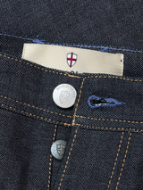 Close-up of Giacomo Selvedge V2 Jeans - Raw Denim by Blue de Gênes, featuring rigid denim, engraved metal buttons with a shield, gold stitching, and a beige label with a red and white cross. Relaxed loose fit. Made in Italy.