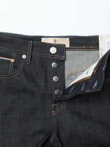 Close-up of Blue de Gênes Giacomo Selvedge V2 Jeans in raw denim: dark blue, rigid, relaxed loose fit with a partially open fly showing four metal buttons, inner lining, yellow stitching, and a white waistband patch with red emblem.