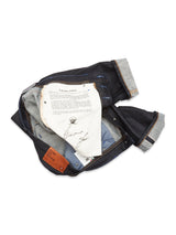 A pair of Giacomo Selvedge Jeans by Blue de Gênes exemplifies Italian craftsmanship with its raw denim, exposed lining, and sewn-in patches. A white document page with text and an autograph accompanies the jeans.