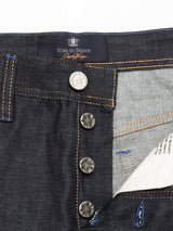 A close-up of raw denim Giacomo Selvedge Jeans displays a partially open button fly, revealing white fabric beneath. The Blue de Gênes brand label stands out inside the waistband above the buttons, and orange stitching accents the rigid denim, highlighting Italian craftsmanship.