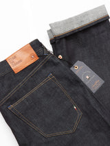 Close-up of Blue de Gênes Giacomo Selvedge Jeans in rigid, dark raw denim with a brown leather waistband patch and visible stitching. The folded cuff reveals a lighter gray interior, highlighting Italian craftsmanship. A branded tag with a small flag detail is attached near the pocket.
