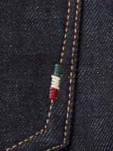 A close-up of dark, rigid denim fabric with orange stitching along a seam highlights the Italian craftsmanship of Blue de Gênes Giacomo Selvedge Jeans - Raw Denim. A small detail features three vertical stripes in green, white, and red embroidered near the seam.