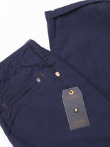 The Lori Tarlo Chino by Blue de Gênes, in navy blue cotton with beige and orange stitching and a black tag featuring a decorative crest, complements its relaxed fit. The pants are neatly folded, showcasing their casual style.