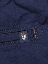 A close-up of the Lori Tarlo Chino by Blue de Gênes shows navy cotton fabric with a small tag. The tag displays sdg in orange letters above a white cross on black. The relaxed fit garments edge is stitched with brown thread, visible near the tag.