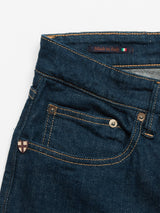 Close-up of blue denim jeans highlighting the front pocket, button, and a small cross badge. Above is a labeled Blue de Gênes crafted in Italy with an Italian flag design. Classic orange stitching on Comfort Stretch Denim offers style and ease for Vinci Topaz Jeans - Rinse Wash.