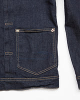 The Cosimo Trucker Jacket - Dark Blue Denim by Blue de Gênes is detailed with a small, buttoned front pocket accented by light stitching, a metal rivet, and subtle embroidery. Its fabric presents a slightly textured look.