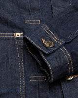 A close-up of the Blue de Gênes Cosimo Trucker Jacket in dark blue denim highlights visible orange stitching. Focus is on the cuff with a metal button featuring engraved text, alongside a partially visible pocket with a metal rivet, emphasizing the jackets western-inspired texture.