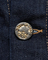 The close-up shows a silver button on the Cosimo Trucker Jacket in Dark Blue Denim by Blue de Gênes, featuring Blue de Gênes and a crown design. Light brown stitching around the buttonhole enhances its stylish durability.