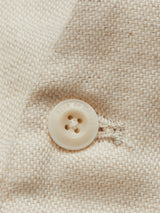 Close-up of the Pantella Desk Gilet - Natural by Blue de Gênes, showcasing its beige, textured Cotton/Linen blend fabric with a woven pattern. A beige Corozo button with engraved text provides a subtle contrast to the fabrics texture.