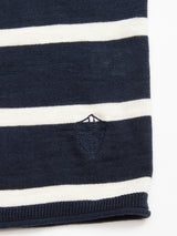 A close-up of the Monte Stripe Knit - Navy by Blue de Gênes shows a navy and white horizontal striped fabric with a small embroidered shield emblem. Made from a soft cotton/linen blend, it resembles a classic crewneck jumper, with the fabric appearing folded at the bottom edge.