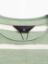 Close-up of a Monte Stripe Knit - Green Bay jumper from Blue de Gênes, featuring a label with emblem inside the collar. Made from a cotton/linen blend, the soft knit fabric displays green and cream stripes.