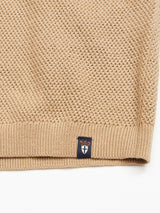 A close-up of the Filo Knit Polo - Khaki by Blue de Gênes reveals a beige textured 100% cotton knit fabric with a waffle-like pattern. A small black tag featuring a white shield and orange text enhances the retro repeat stitch design at the bottom edge.